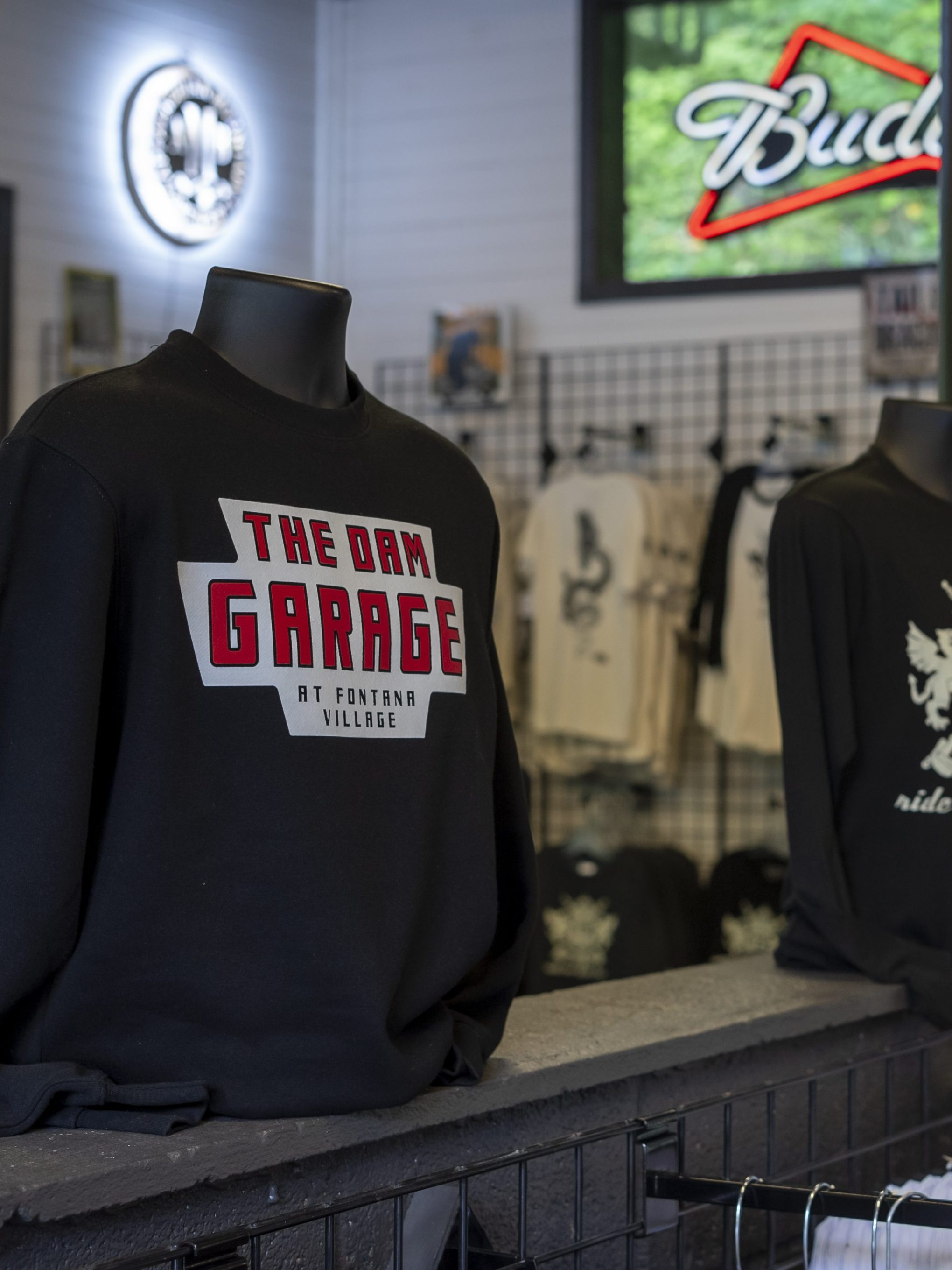 Mannequins display black sweatshirts in a shop, with snacks and a neon sign in the background.