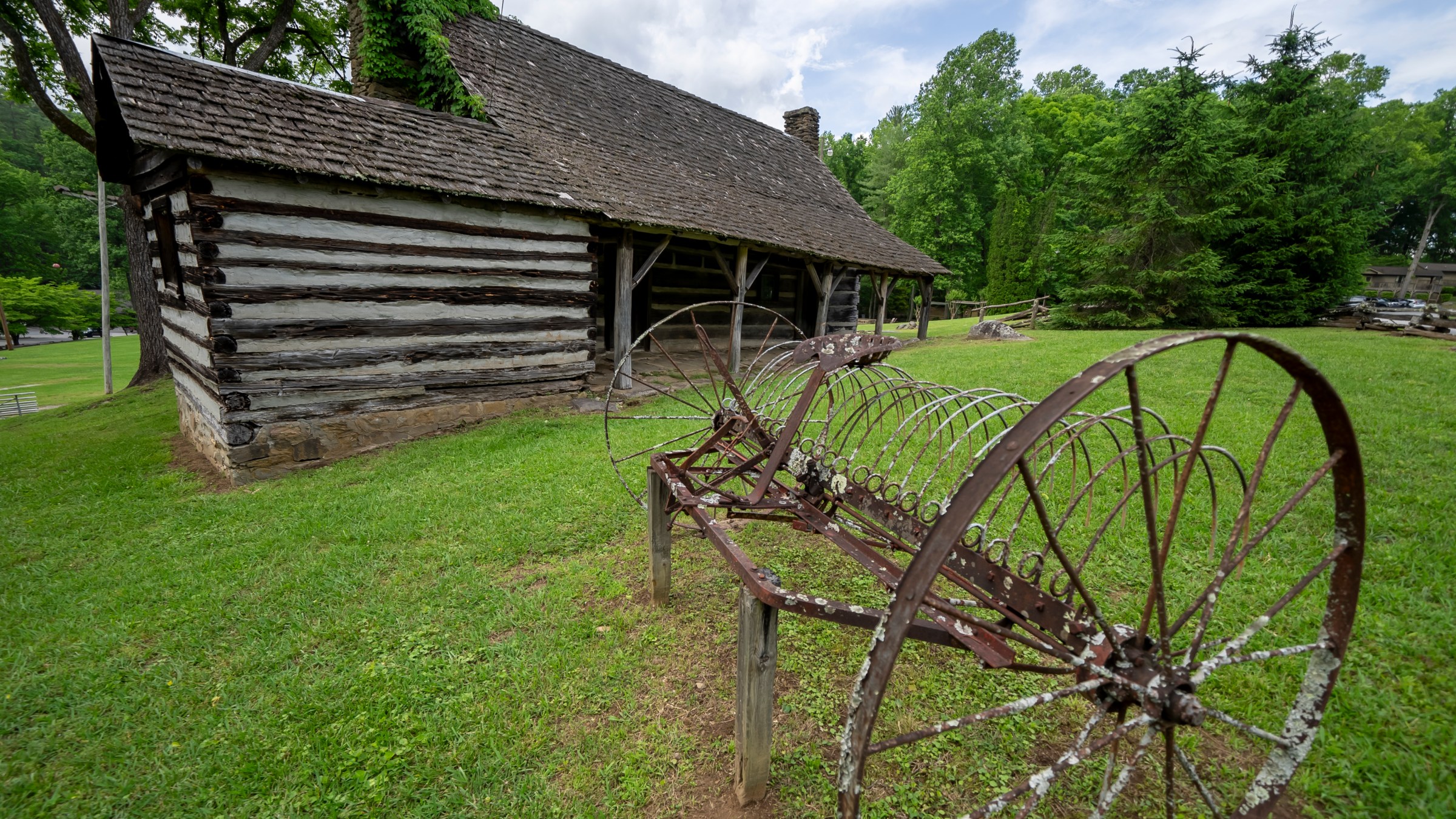 Old wooden cabin with a rusted metal farming tool on grassy field, surrounded by trees.