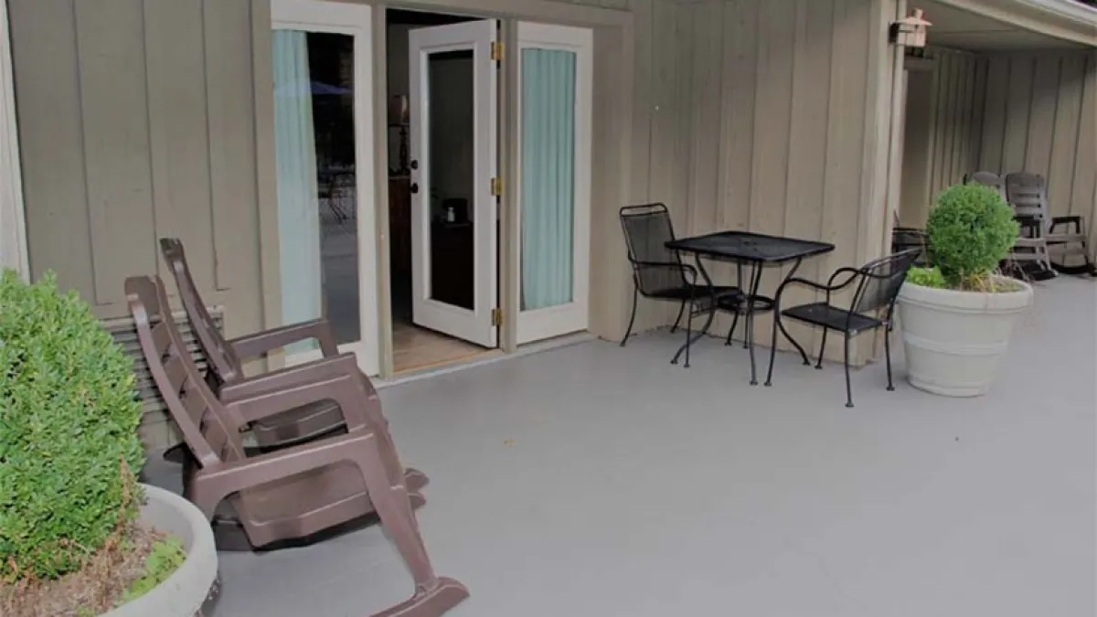 Patio with two rocking chairs, a table with chairs, and potted plants outside a building's open doors.