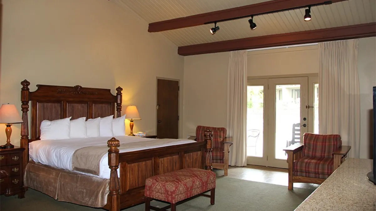 Spacious hotel room with a large wooden bed, armchairs, and glass doors leading outside.