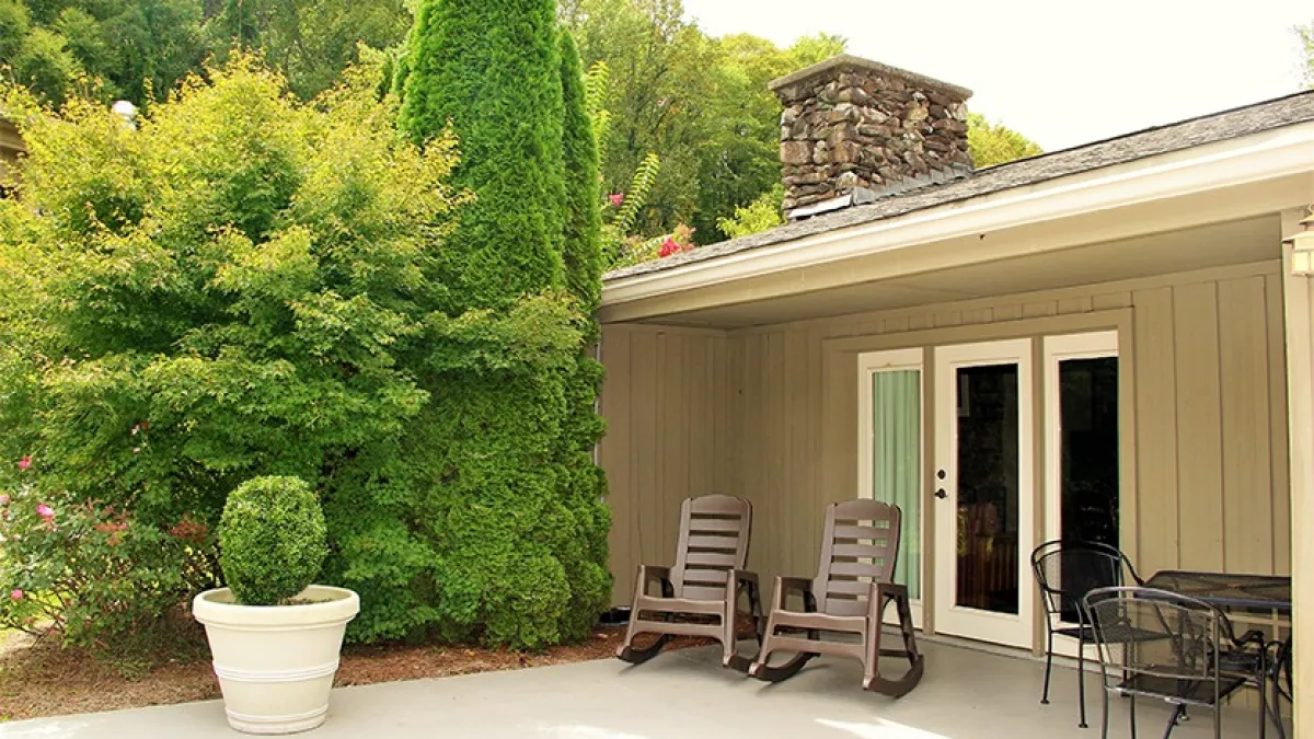 Patio with two chairs, table set, potted plant, and greenery by house entrance.