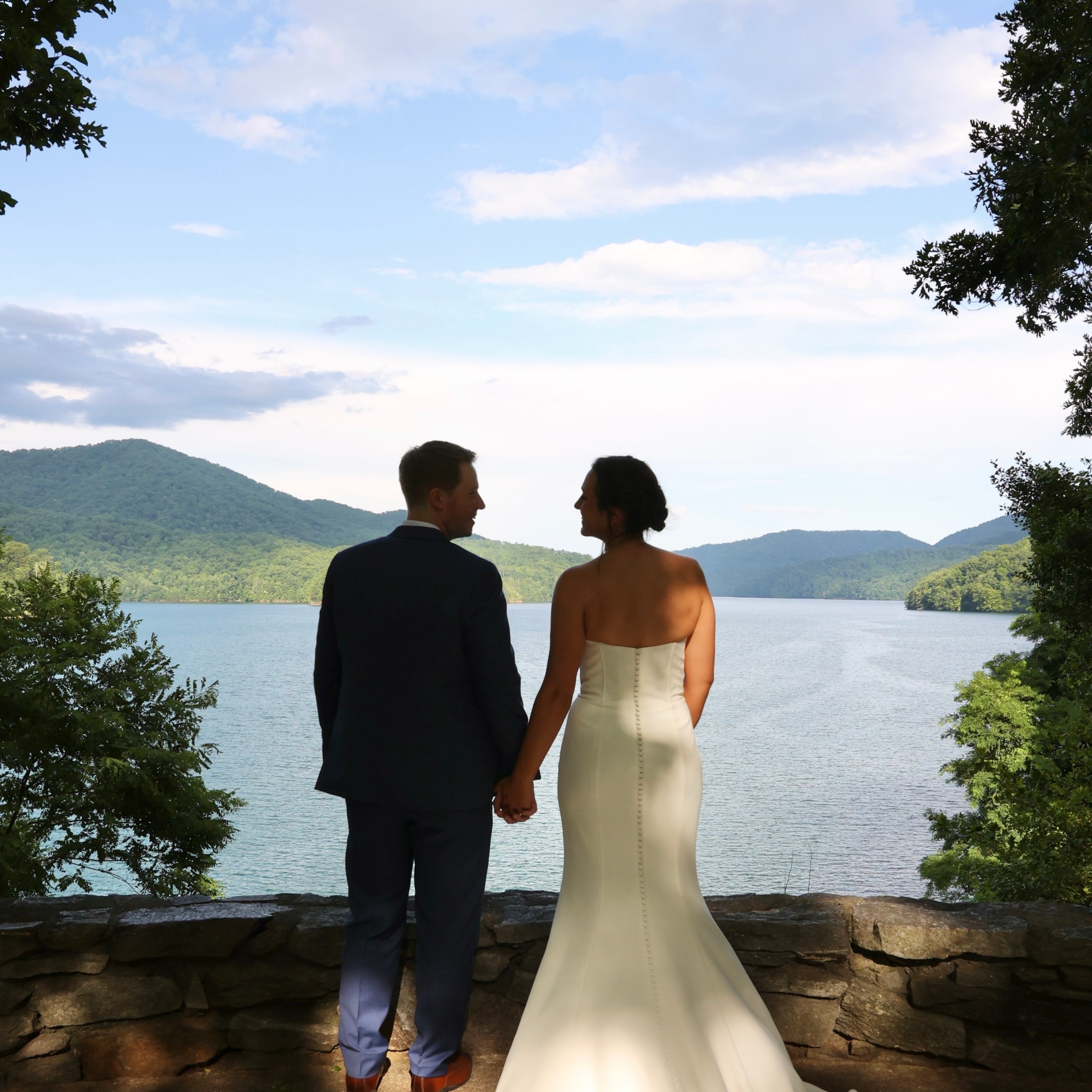 Couple in wedding attire holding hands, overlooking a scenic lake and mountains.