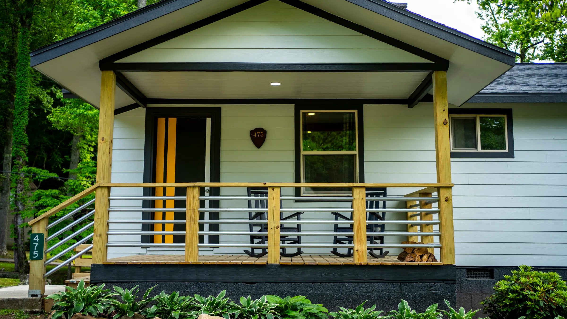 Small house with porch, wooden railing, and striped door, surrounded by trees and plants.