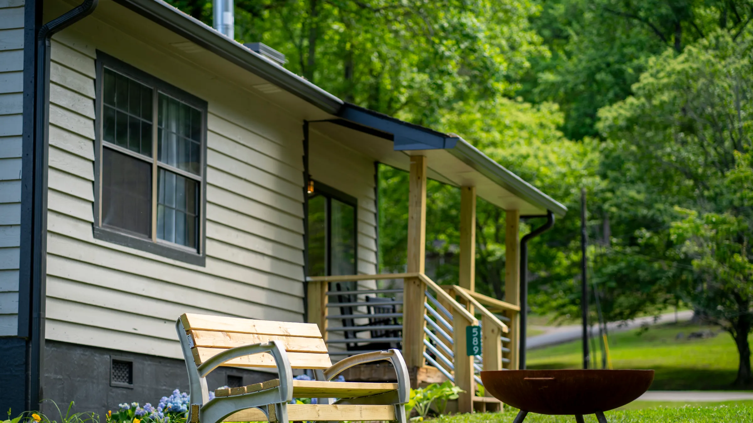 House with wooden bench and fire pit in grassy yard, surrounded by trees.