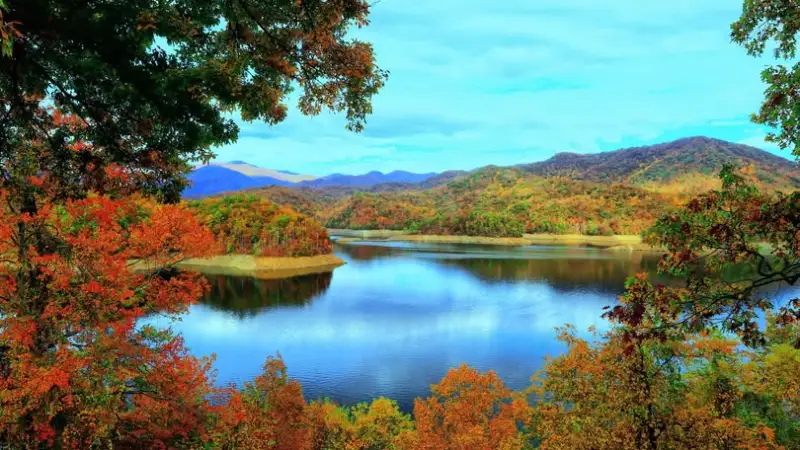 Lake surrounded by colorful autumn trees and distant hills under a partly cloudy sky.