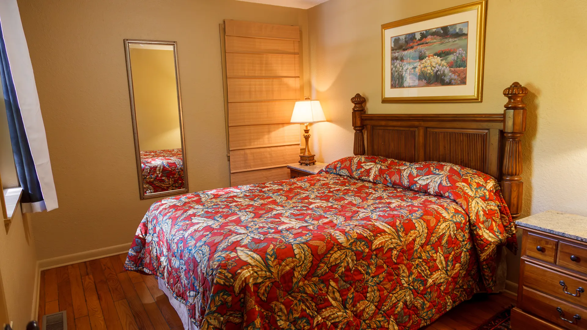 Cozy bedroom with floral bedspread, wooden headboard, lamp, and wall art. Hardwood floor and mirrored closet.