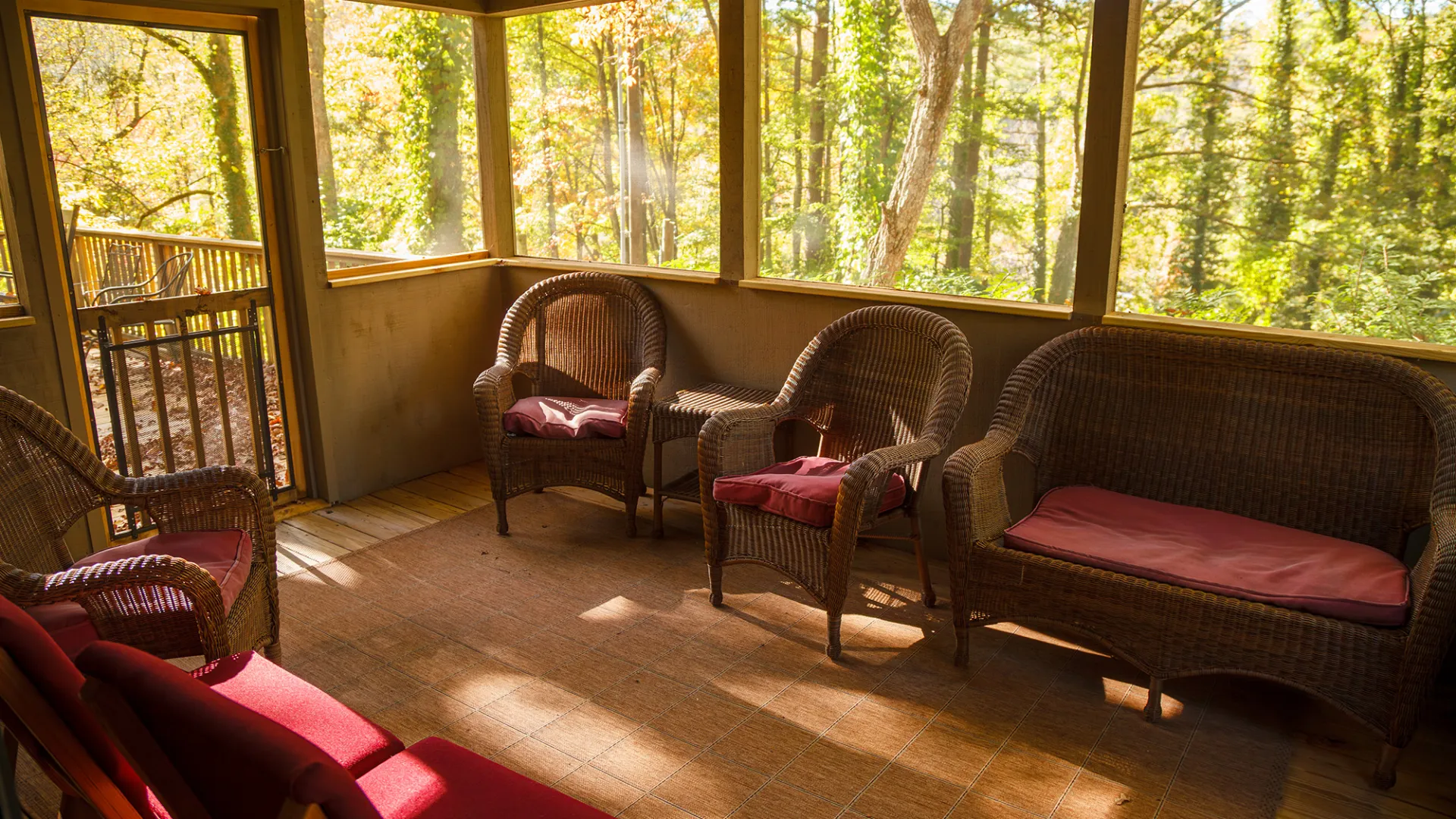 Sunlit porch with wicker furniture and red cushions, surrounded by trees.