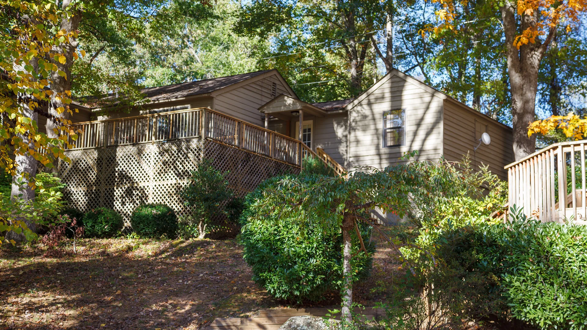 A brown house with a wooden deck surrounded by trees and bushes on a sunny day.