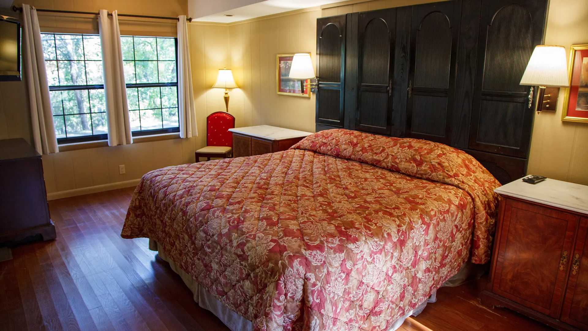 Cozy bedroom with red quilted bedspread, wood floor, lamps, and large windows with curtains.