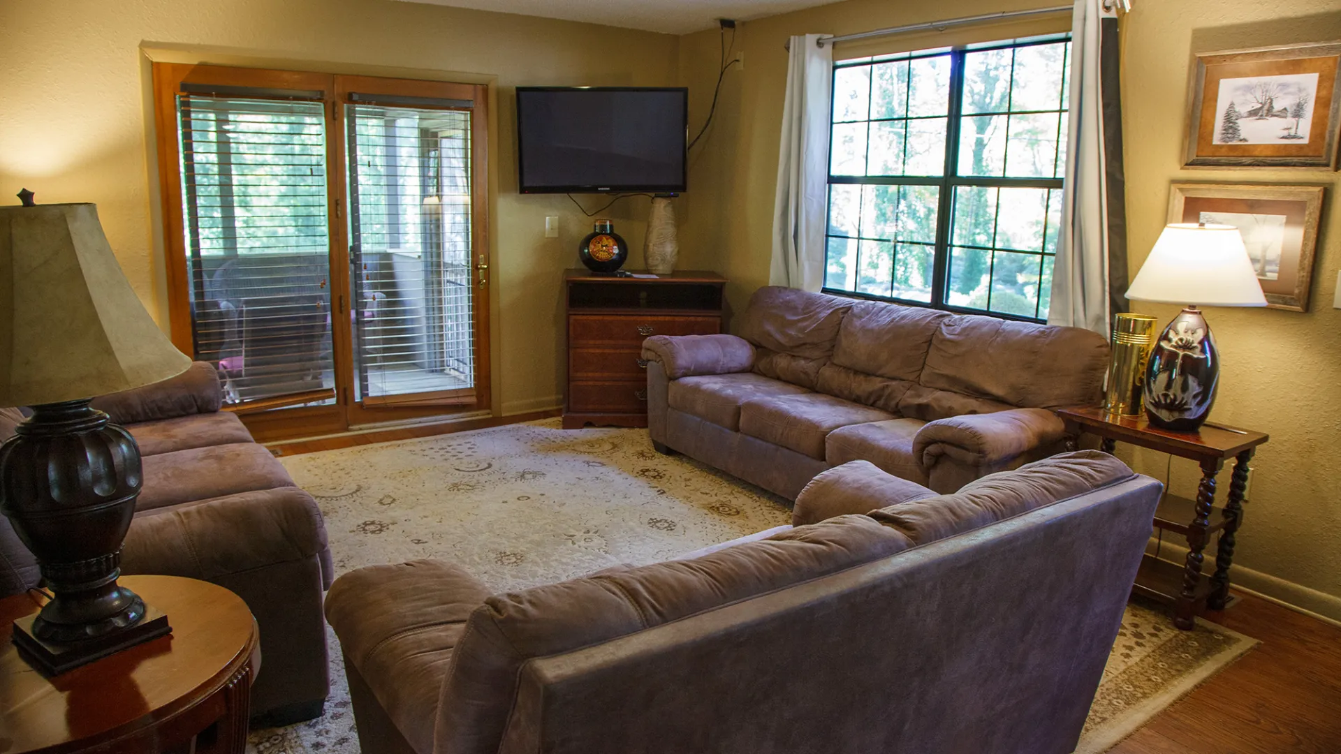 Cozy living room with brown sofas, rug, lamps, TV, and window with curtains facing a door to a sunroom.
