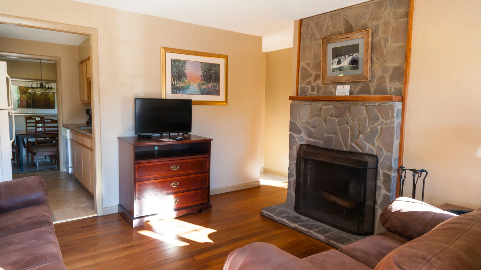 Living room with stone fireplace, TV on dresser, and couches, leading to kitchen.