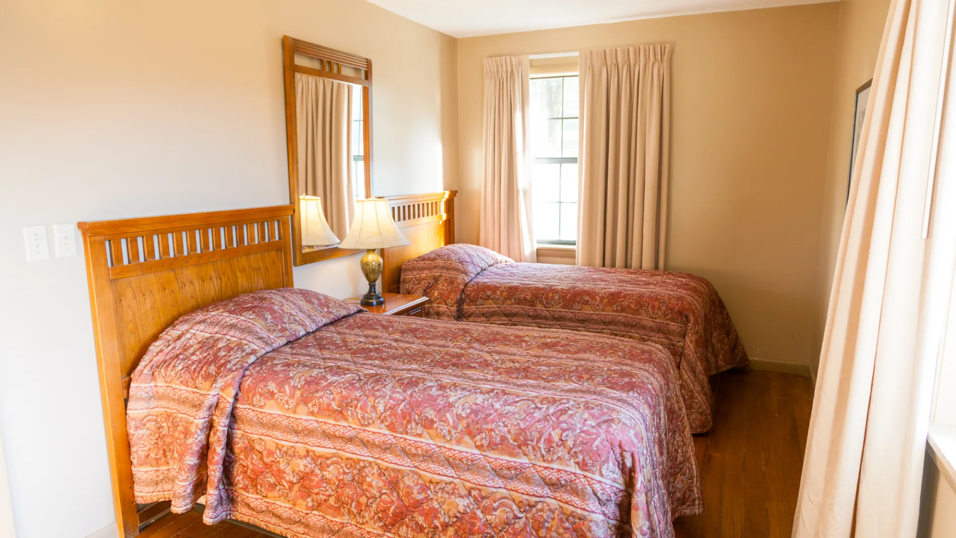 Bedroom with two wooden beds and patterned bedspreads, nightstand, lamp, and window with curtains.