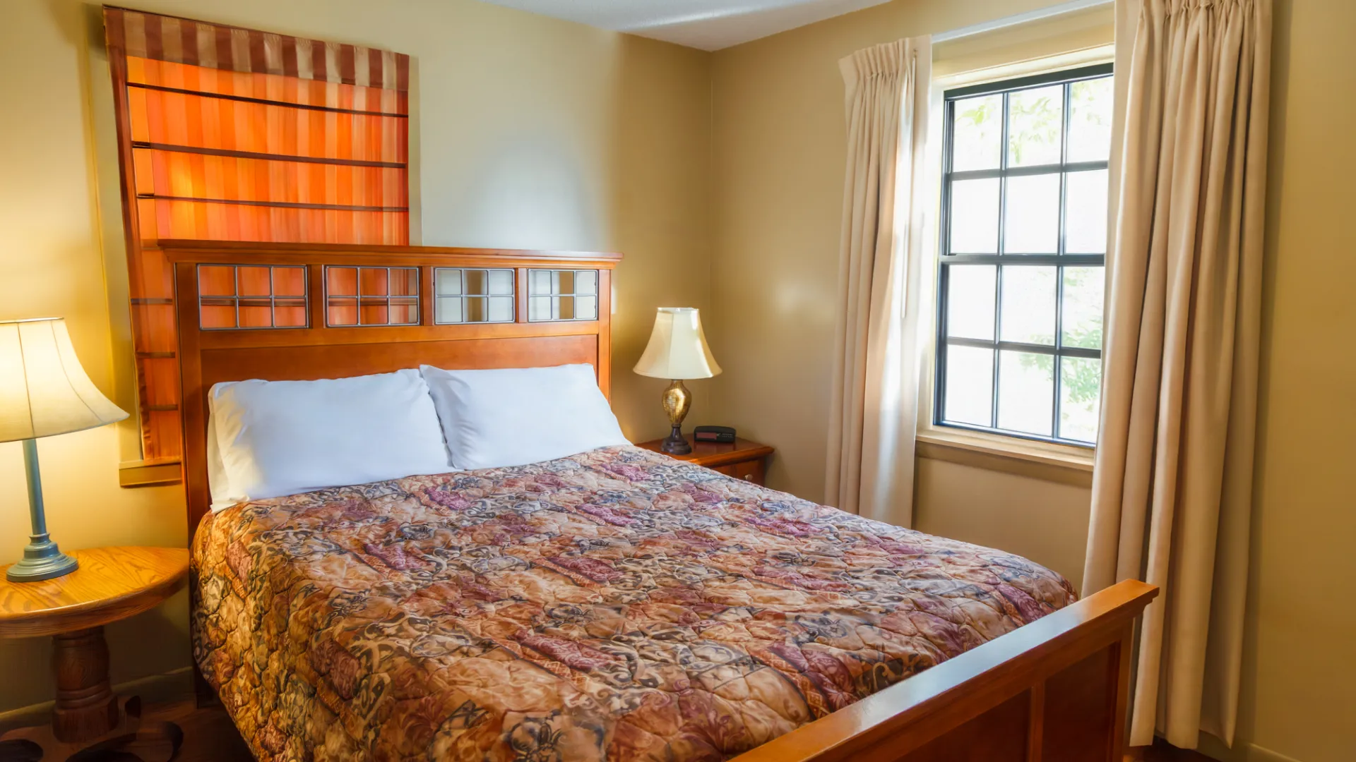 Cozy bedroom with a wooden bed, patterned quilt, lamps, and a window with curtains.