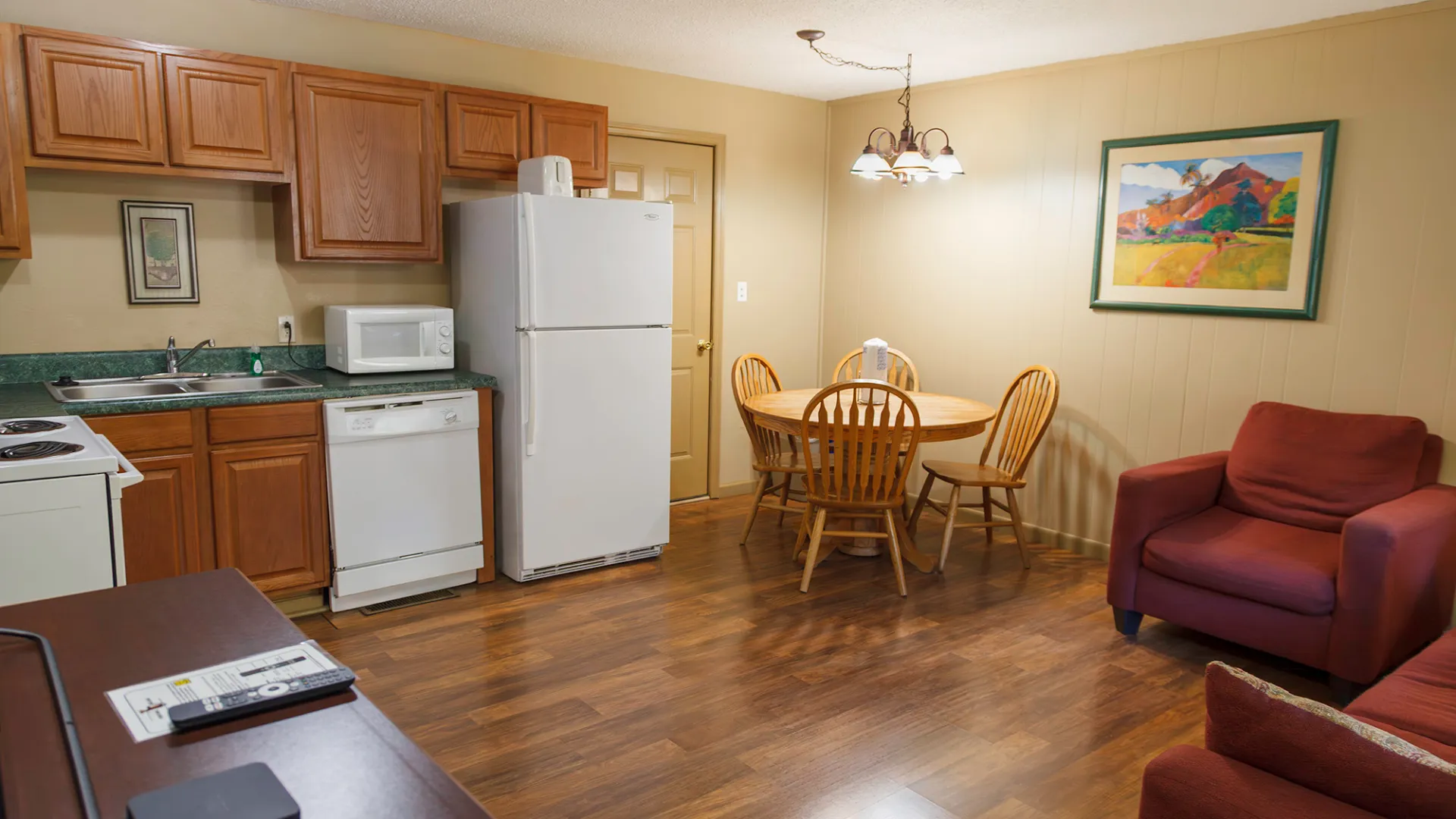 Small kitchen with wooden cabinets, dining table, chairs, fridge, armchair, and wall art.