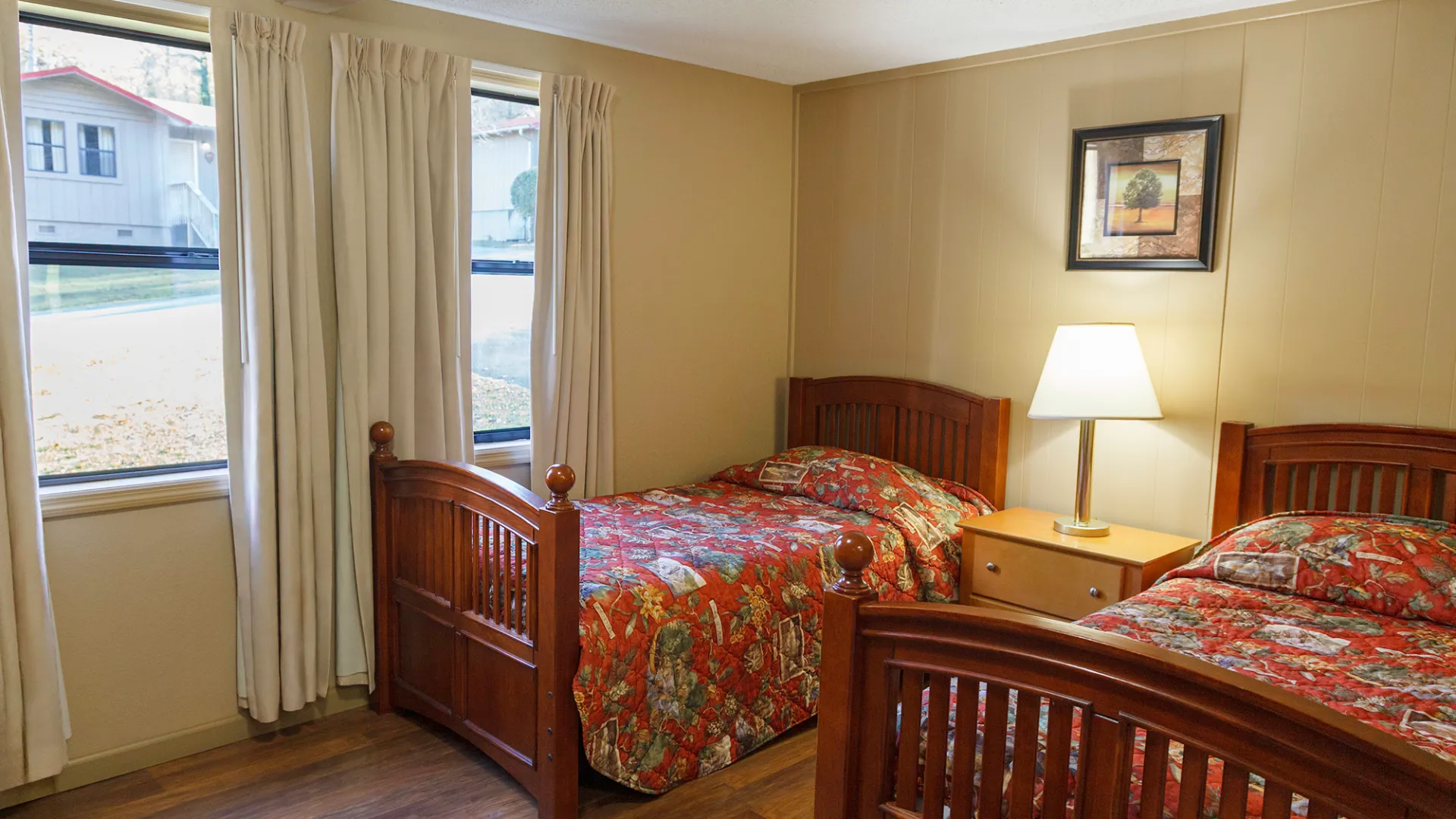 Room with two wooden beds, colorful bedding, a lamp on a nightstand, and a picture on the wall.