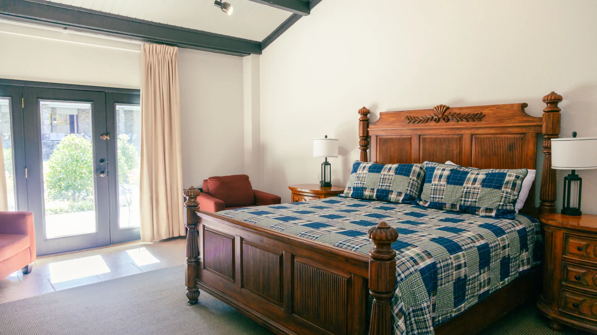 Bedroom with wooden bed, plaid bedding, nightstands, lamps, armchair, and glass doors.