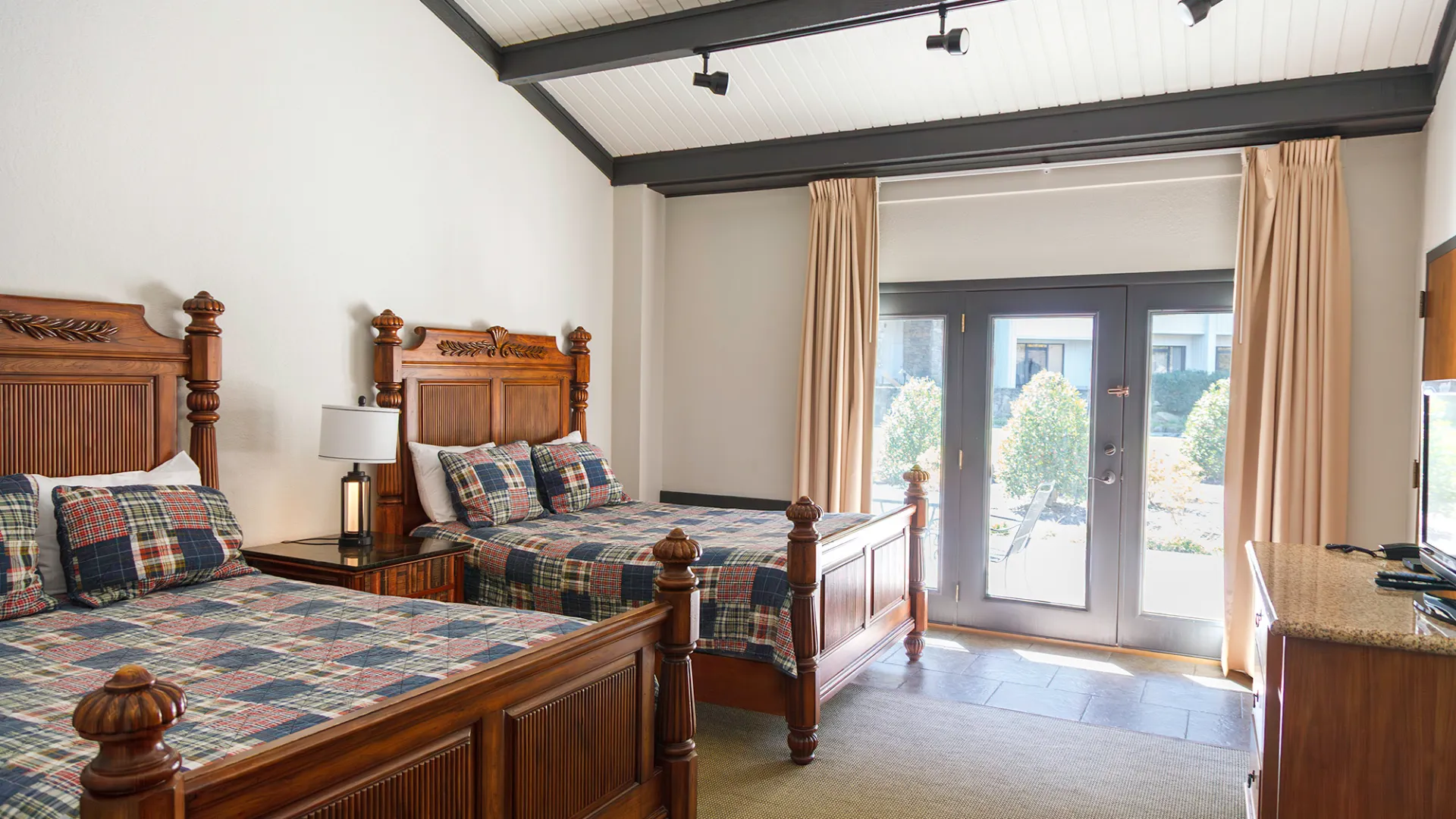 Room with two wooden beds, plaid bedding, lamp on nightstand, and glass doors with beige curtains.