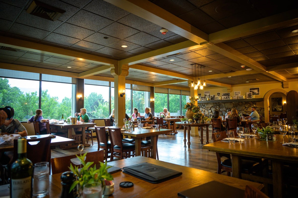 Spacious restaurant with wooden tables, large windows, and diners seated. Greenery visible outside.