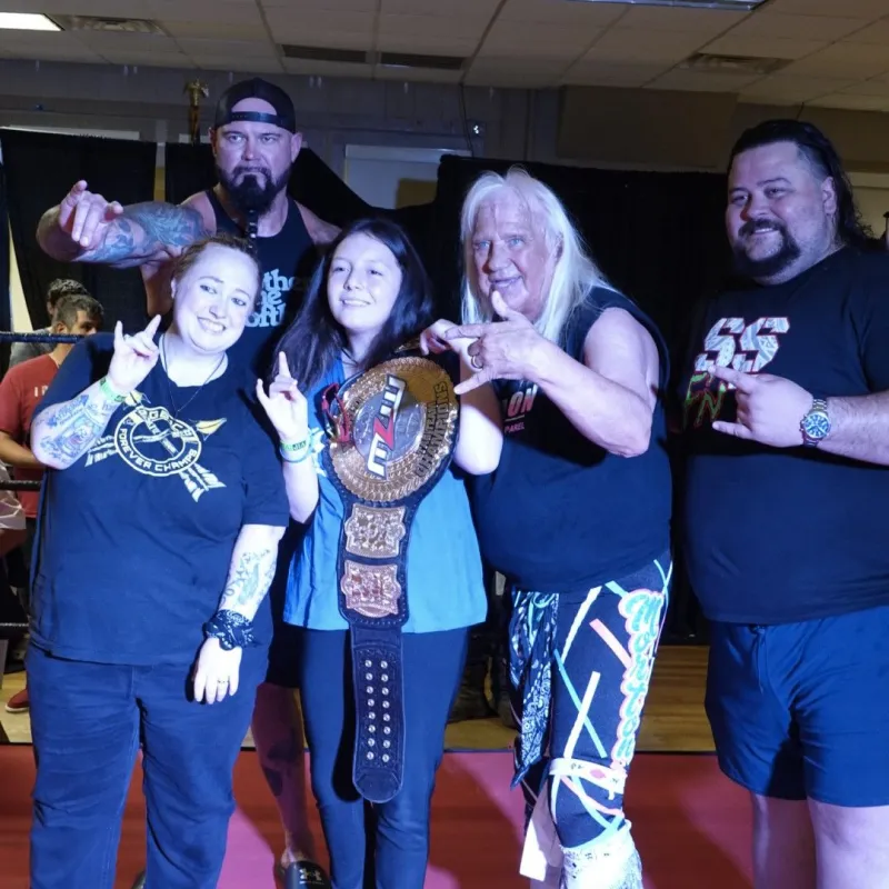 Group of five people, one holding a wrestling belt, posing together indoors.