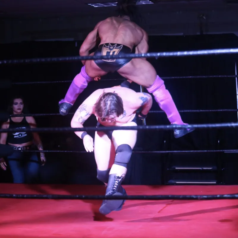 Wrestler jumps over opponent in a wrestling ring, referee nearby.